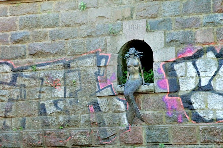 Seduta in una nicchia sopra il fiume, la sirena guarda verso il cielo (Uzupis Mermaid- Lithuania